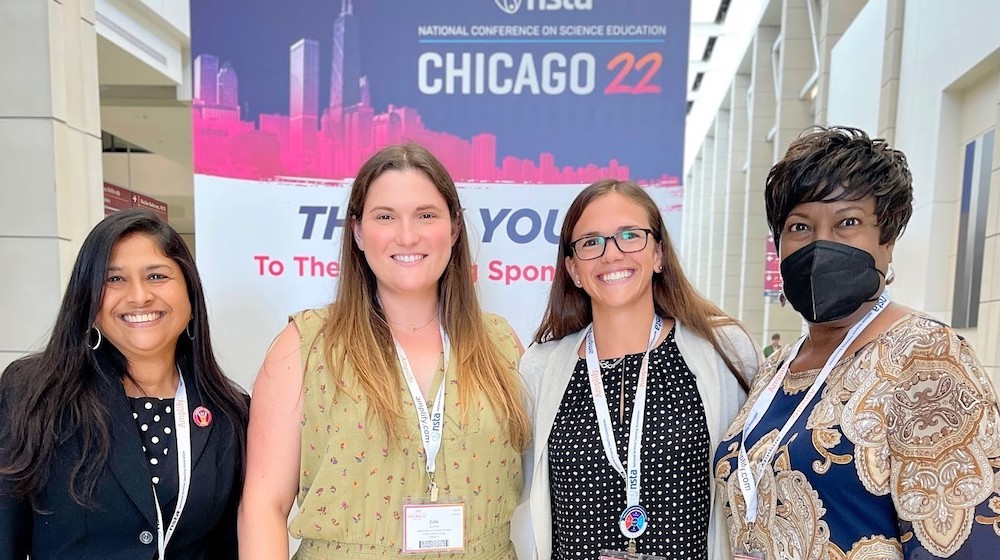 Four women, all Loyola University Chicago CSME representatives, stand in front of the National Science Teaching Association Conference banner.