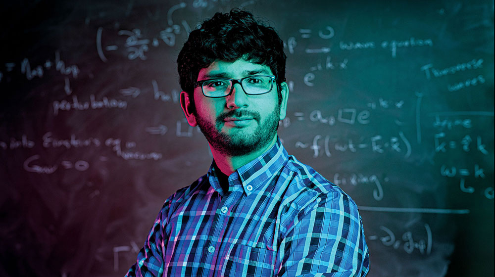 Headshot of Loyola University Chicago physics professor Walter Tangarife sitting in front of a blackboard with chalk equations written on it.