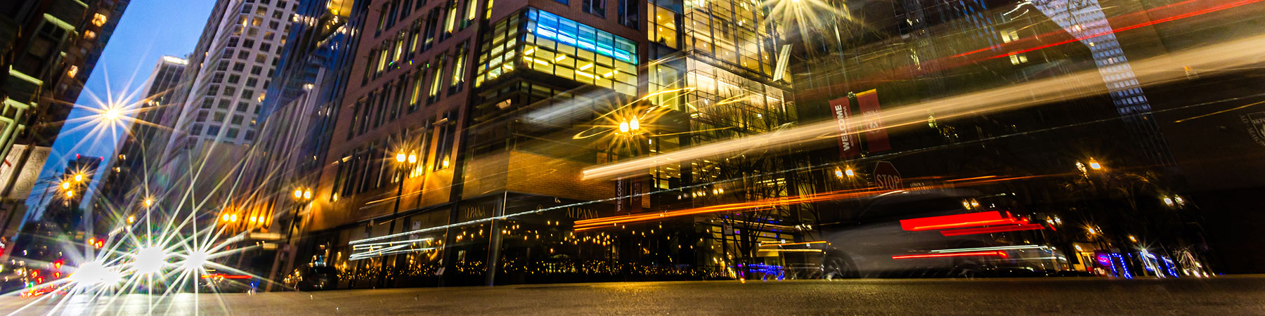 The evening sky reflects in the windows of Water Tower campus buildings at dusk while streaking red and gold vehicle lights and commuters pass in the foreground.