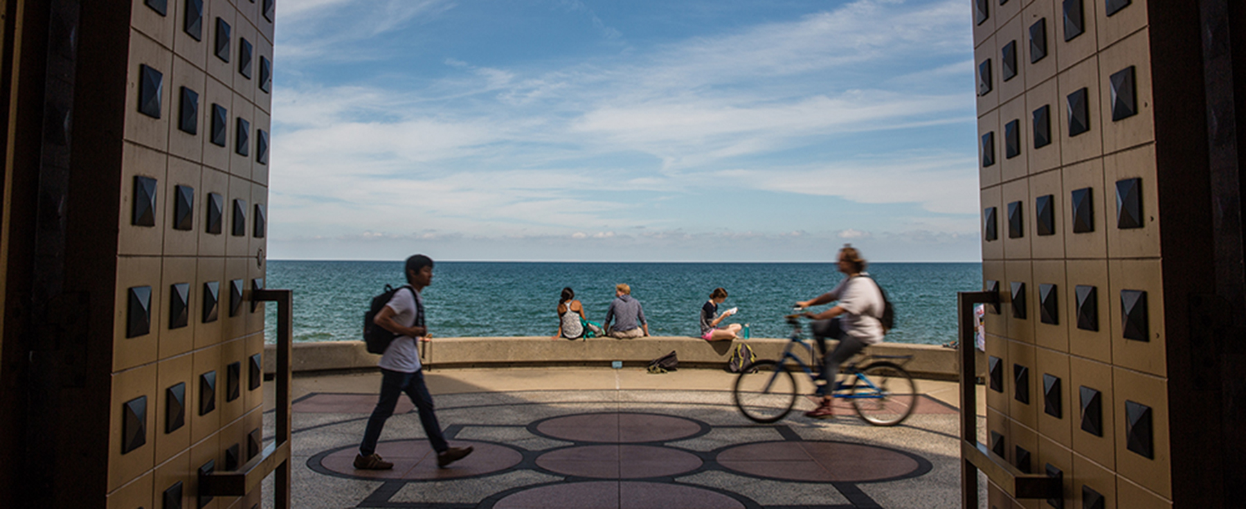 Taken from inside the front doors of Loyola University Chicago's Madonna della Strada Chapel on the Lake Shore Campus. It is a sunny day with a few clouds, overlooking Lake Michigan. Students are walking by, riding a bike by, and a few are sitting on the retaining wall on the edge of the water. 