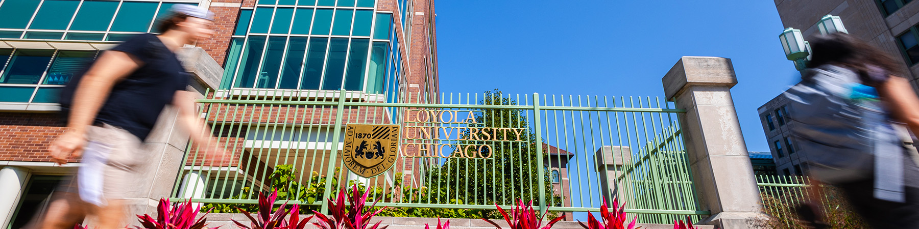  Students in motion, walking past Loyola University Chicago's metal logo signage on the Lake Shore Campus. It's a sunny day with blue skies and red brick campus buildings in the background.