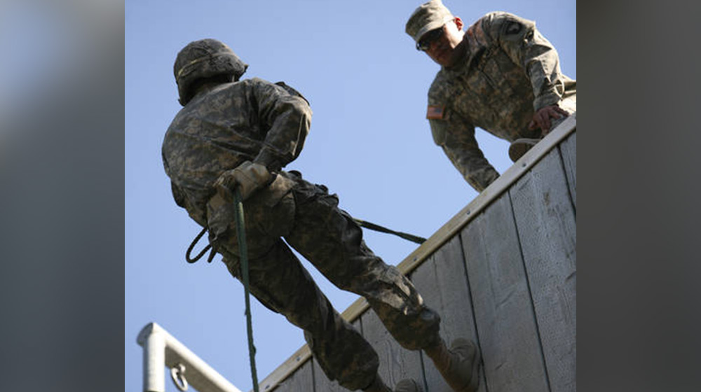 A military trainee in uniform rappels down a wall while a instructor watches from above