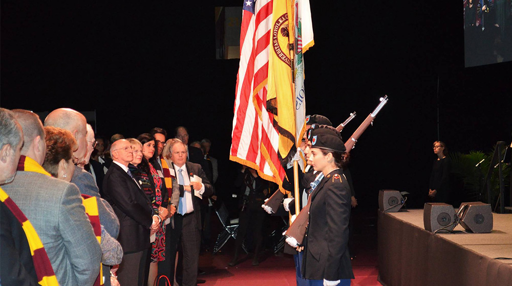 A color guard presents the U.S. flag, another flag, and the ceremonial rifles at an indoor event. Audience members, some in formal attire, stand at attention, and a person in a black robe is seen on stage in the background.