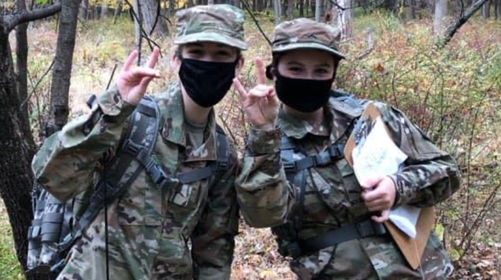 Two cadets in military uniform pose for a photo together in a forest. They are both wearing masks and making hand symbols resembling a wolf. One is holding a clipboard.