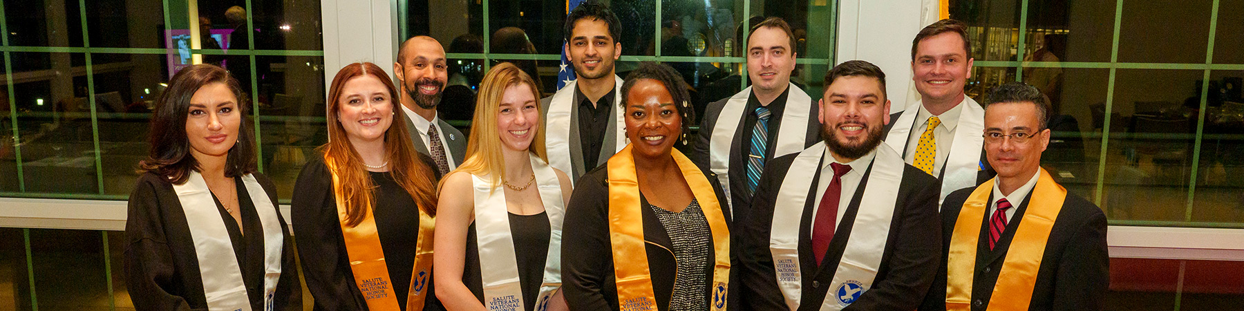 A group of ten people from the Loyola Military Veteran Honors Society stand in two rows, smiling at the camera. They are dressed in formal attire, and several are wearing honorary sashes. The background shows large windows with a nighttime cityscape visible through them.