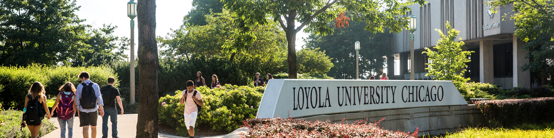 Students walk the campus of Loyola University Chicago in the spring. There is a large stone sign that has the name of the university on it.