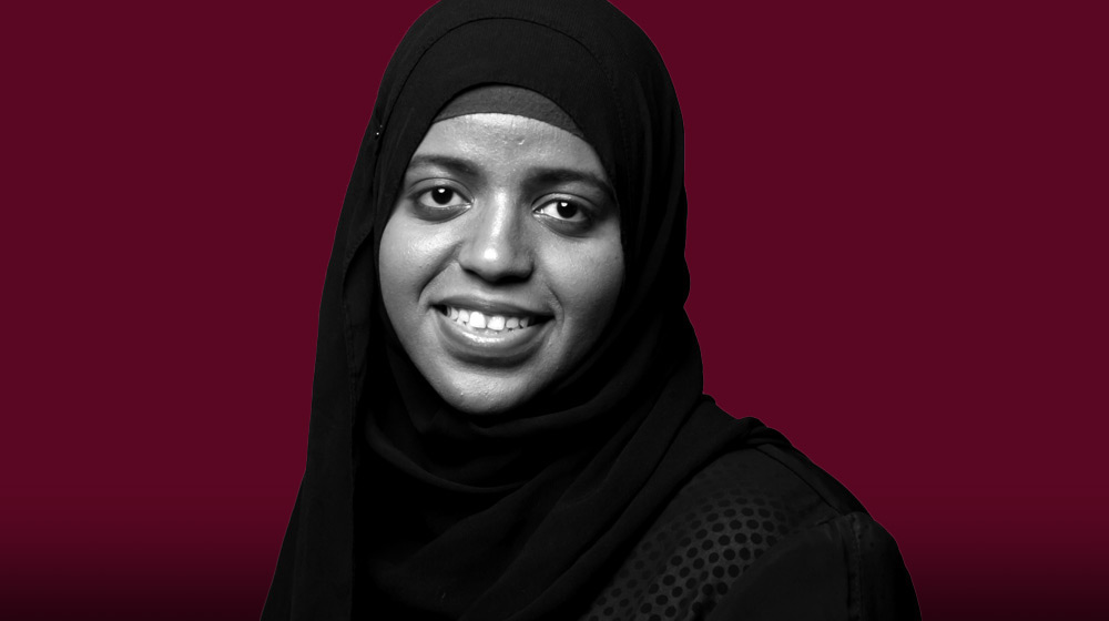 A black and white portrait of a Loyola University Chicago nursing student in a hijab, smiling. They are centered against a maroon background.