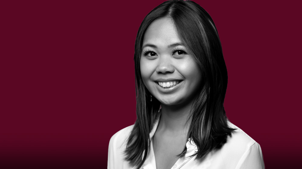 A woman with shoulder-length hair smiles while looking at the camera. She is wearing a white blouse and is positioned against a maroon background.