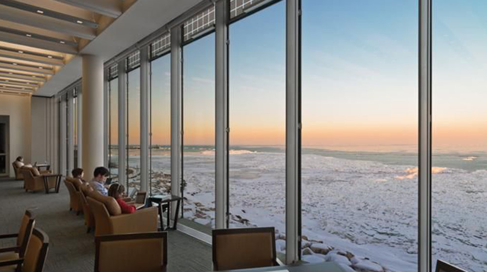 Multiple students sit in large leather chairs overlooking a frozen lake from floor to ceiling glass windows at the Loyola University Chicago Information Commons library