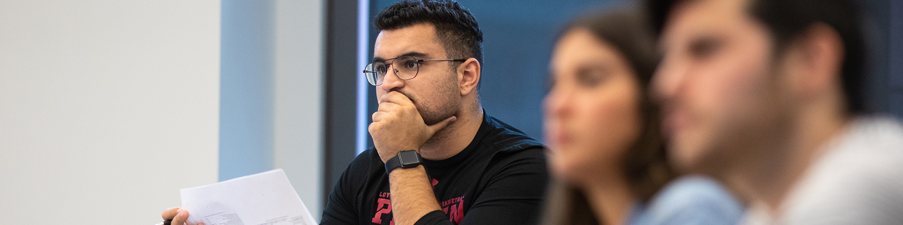 A graduate student with dark wavy hair and glasses, rests his hand on his chin as listens intently to the instructor.